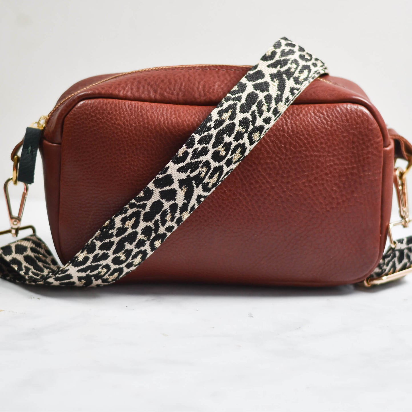 Brown leather handbag with a leopard print strap on a light gray background
