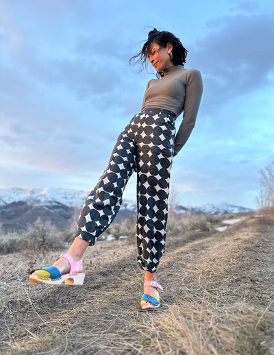 Woman in patterned pants and colorful sandals standing on a mountain trail with mountains in the background.