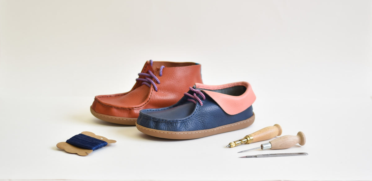 Two handmade leather shoes displayed on a white studio background, one in reddish-brown and one in navy blue with a pink collar, accompanied by leatherworking tools and thread.
