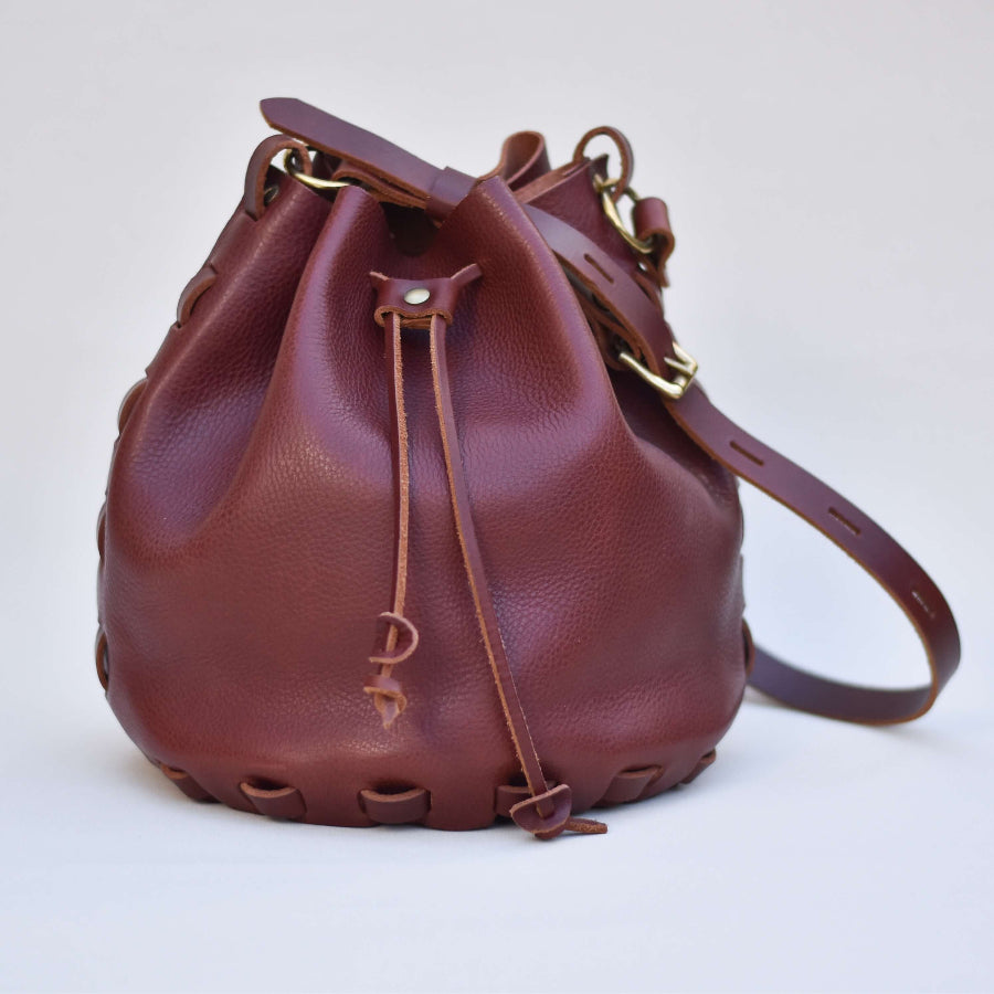 A dark burgandy brown leather bucket bag with a drawstring closure and long strap made from a kit