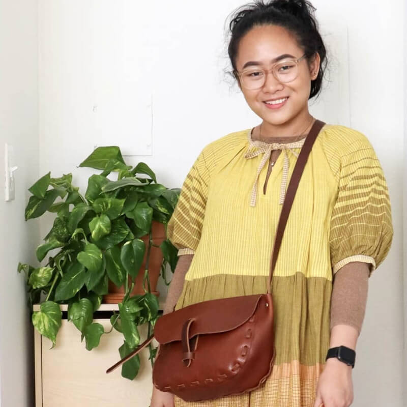 Woman in a yellow dress with a brown bag standing next to a potted plant indoors.