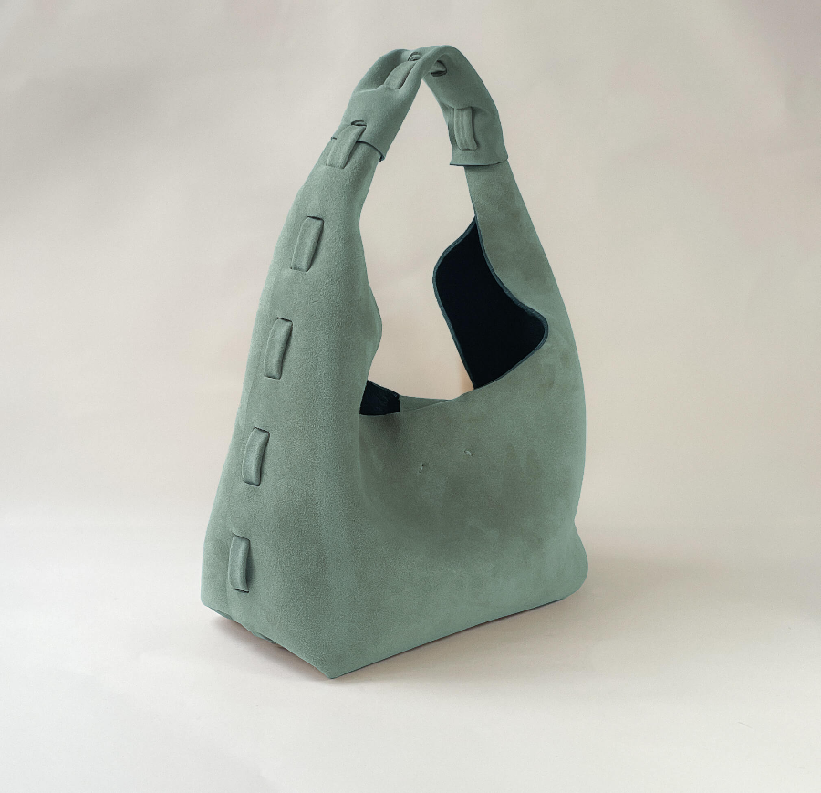A sage green suede shoulder bag with a unique slouchy silhouette, featuring a single wide strap, decorative rectangular stitched details along the side, and a dark navy blue lining visible inside the main compartment, set against a plain light background.