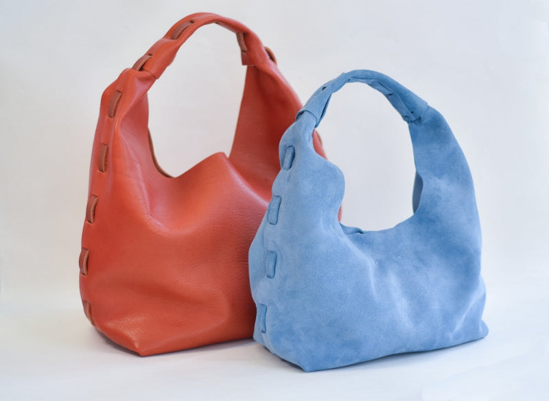 Two slouchy shoulder bags with cutout side details, one in coral orange leather and one in light blue suede, displayed on a clean white background.